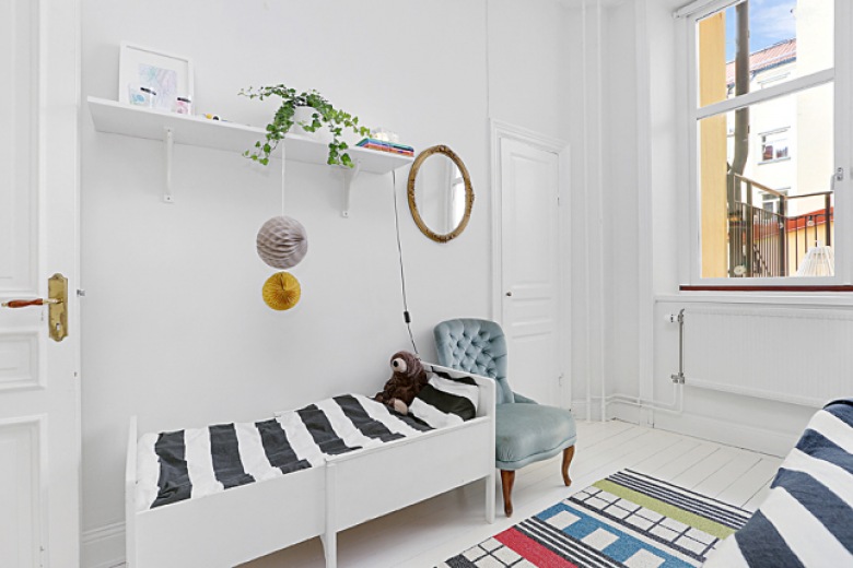 Biało-czarna pościel w paski,białe łóżko z drewna,kolorowy geometryczny dywanik i okrągłe lustro na ścianie w sypialni (26758)