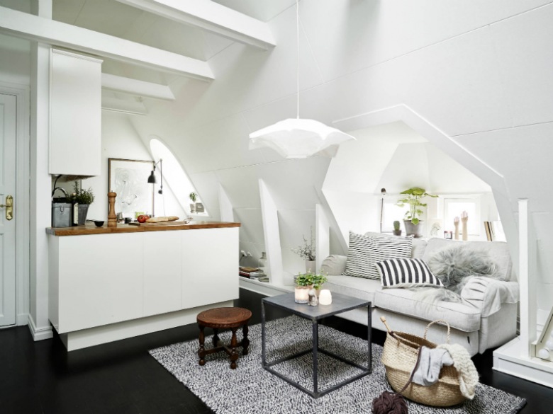 Otwarty salon z kuchnią , białe skośne ściany zbelkami,czarna podłoga,biała sofa na kółkach,metalowy stolik kawowy z drewnianym blatem,szary dywan i biało-czarne poduszki (27318)