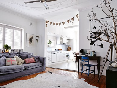 Szara sofa,bydlęca biała skóra na podłodze,niebieskie krzesło,proporczyki i biała kuchnia w otwaretej przestrzeni mieszkania (24755)