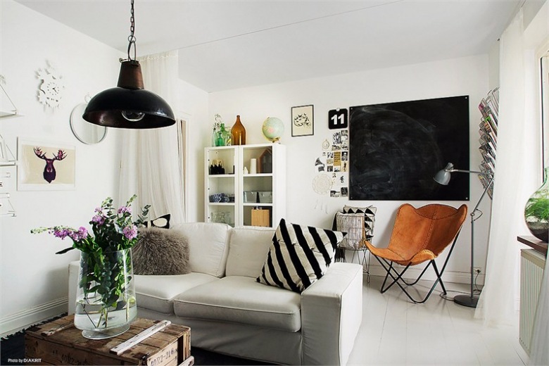 Mały bialy salon z czarną tablicą, fotelem butterfly,lampą industrialną i drewnianą skrzynią (23789)