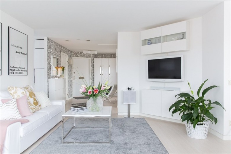 Szary dywan,kwadratowy stolik na chromowanych nogach,biała sofa z różowo-pudrowymi poduszkami w otwartym salonie (28629)