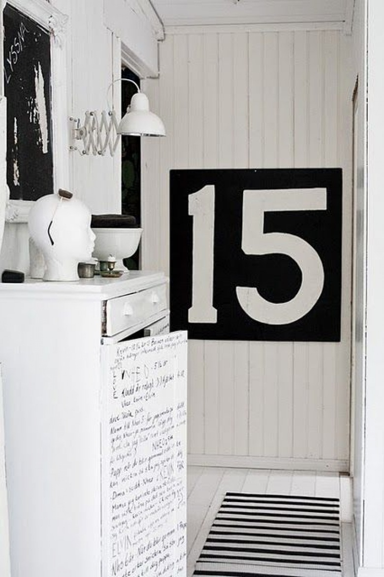 Biała ściana z desek i czarna tablica z cyframi, biała komoda z czarnym nadrukiem pisma odrecznego,bialy metalowy kinkiet z wysięgnikiem i biało-czarny dywanik w paski na białej podłodze (26068)