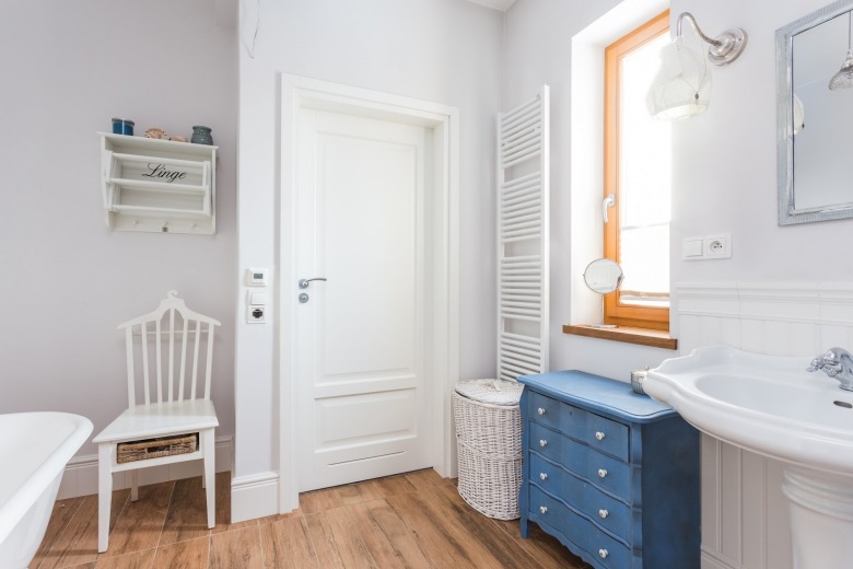 Biała łazienka z niebieskimi dodatkami. Podłoga imitująca drewno oraz wiklinowe dekoracje dodają jej uroku i ocieplają całe...