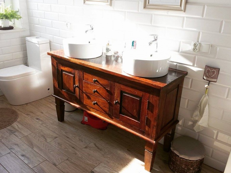 Aranżacja białej łazienki z drewnianą podłogą i rzeźbioną szafką. Dwie umywalki zapewniają wygodny równoległy dostęp...