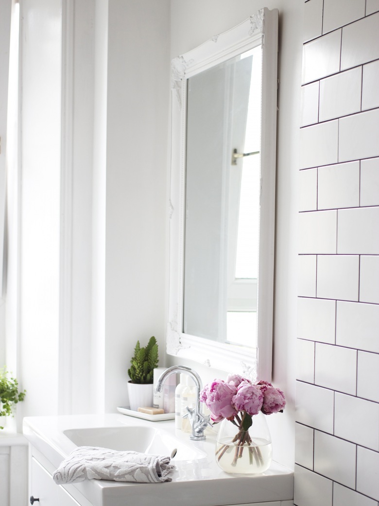 Dekoracyne, białe lustro nad umywalką nadaje aranżacji uroku, dodatkowo podkreślonego różowymi kwaitami w szklanym wazoniku. Białe ściany, sufit, szafka pod umywalką i płytki rozjaśniają wnętrze i sprawiają, że wydaje się higienicznie...