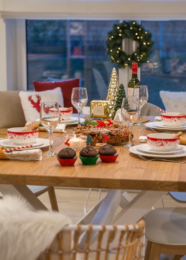 Na drewnianym stole znajduje się kilka dekoracyjnych przedmiotów, które zapowiadają nadchodzące Święta. W tle na...