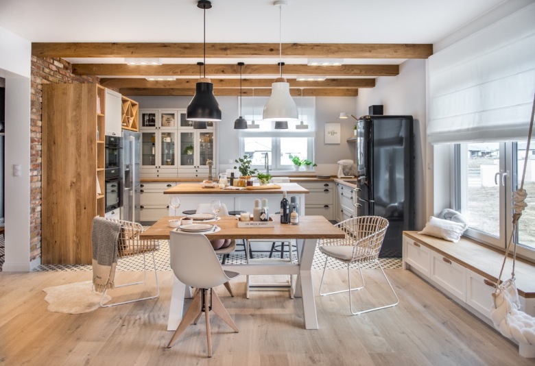 Aranżacja salonu, jadalni oraz kuchni łączy się ze sobą. W otwartej dziennej przestrzeni znajdują się różne drewniane elementy. To nie tylko meble, ale też belki na...