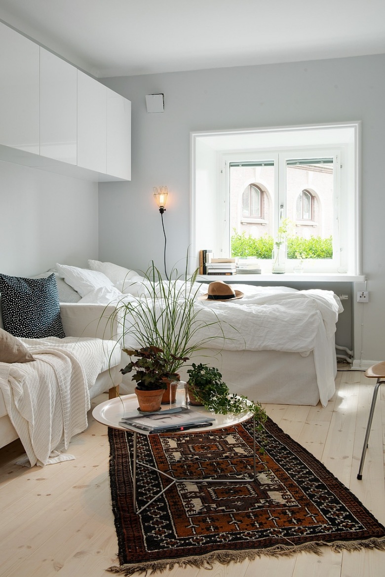 Białe wiszące szafki,łóżko przy oknie i biała sofa, okagłymetalowy stolik z tacą i etniczny brązowy dywan w salonie połączonym z sypialnią (25911)