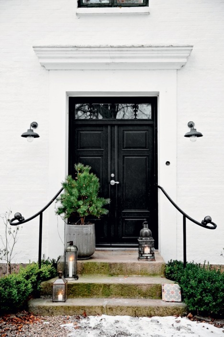 Białe ściany domu,czarne drzwi wejściowe,zielona choinka w szarym wazonie i metalowe latarenki na betonowych schodach (27412)