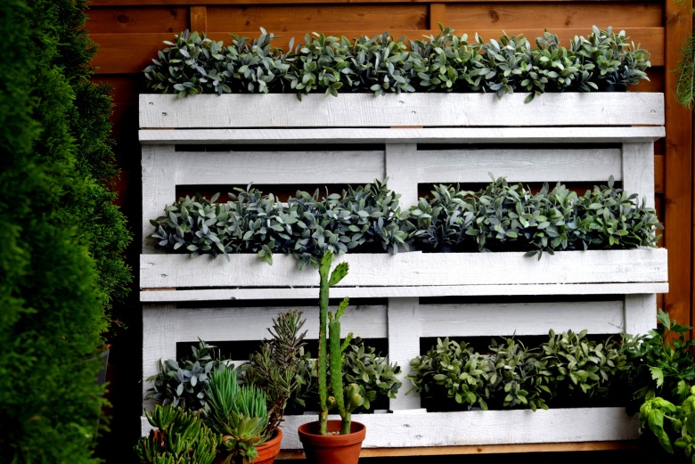 Białą półkę przygotowano z palety, co potęguje rustykalny charakter ogrodowej aranżacji. Zielone rośliny we wdzięczny sposób dekorują praktyczny...