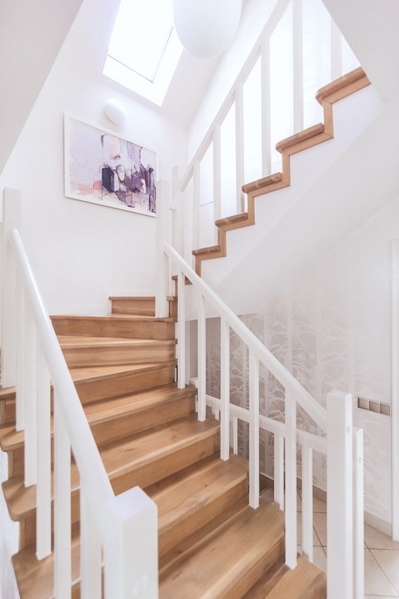 Drewniane schody w białym kolorze rozświetlają przestrzeń. Klatkę schodową znacząco dekoruje abstrakcyjny obraz, który...