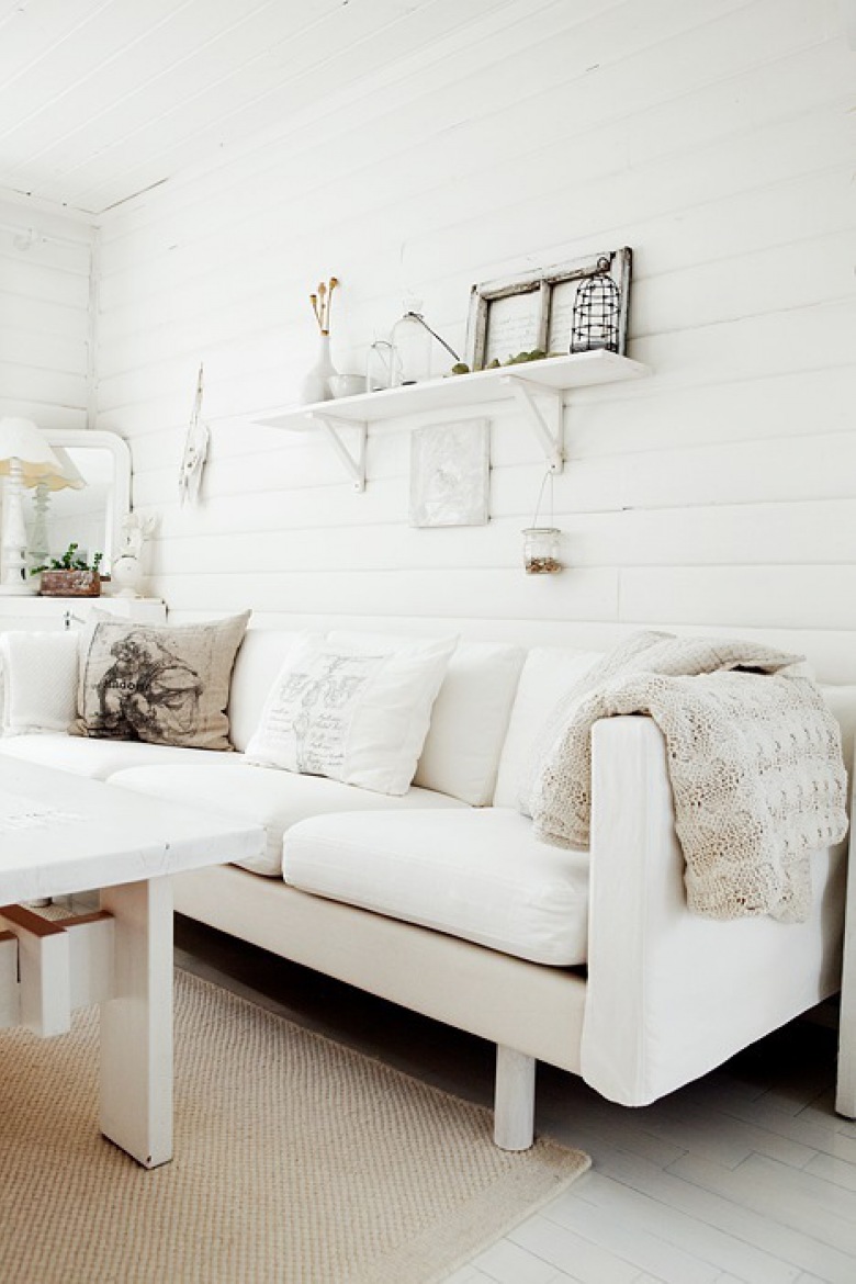 Skandynawska biała kanapa, białe drewniane pólki na scianie i biały drewniany stół w wiejskim salonie (25584)