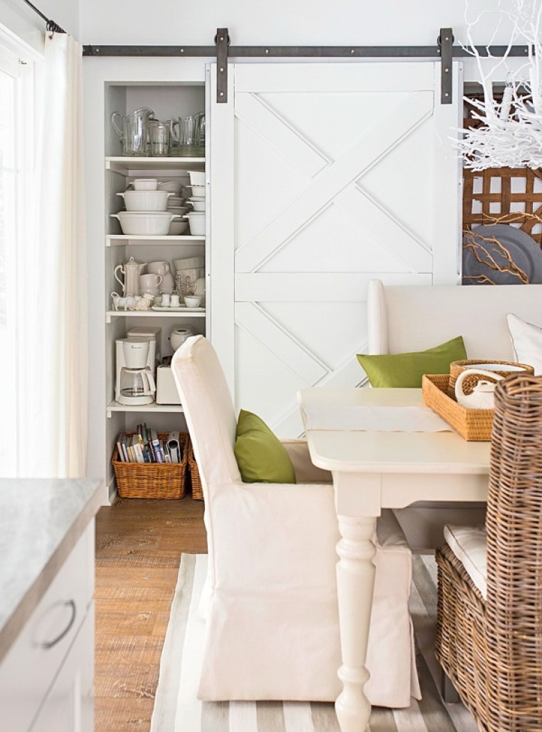 Białe drzwi wrota na żelaznych szynach przy białym regale z półkami w aranżacji jadalni z białym stołem,krzesłami w sukienkach i wiklinowym krzesłem z poduchą (26157)