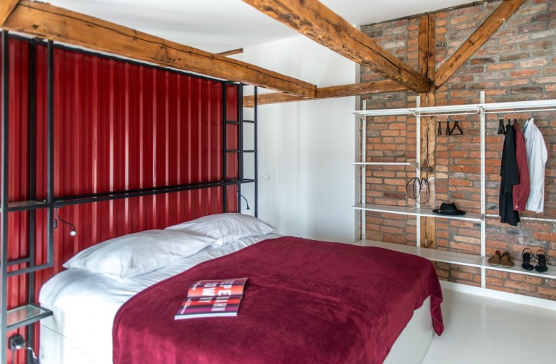 Na ścianie z czerwonymi cegłami w sypialni zorganizowano małą garderobę. Tworzą ją białe wieszaki i półki, które stylem przypominają klimat nowoczesnej lub industrialnej...