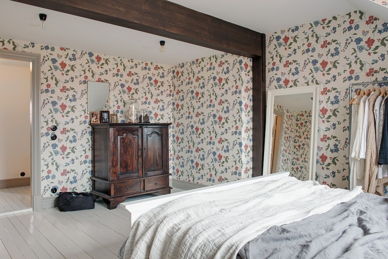 Drewniane ciemnobrązowe belki w sypialni,kwiatowa tapeta ścienna w niebiesko-czerwonym kolorze,retro komoda bieliźniarka przy ścianie,duże białe lustro stojące (25942)