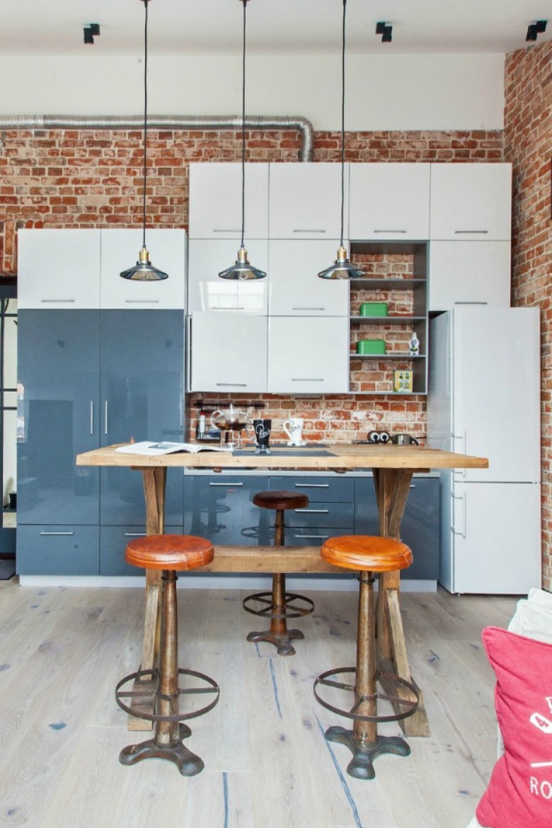 Biało-niebieska kuchnia nowoczesna z industrialnymi lampami,drewnianym stołem i ścianami z czerwonych cegieł (24072)