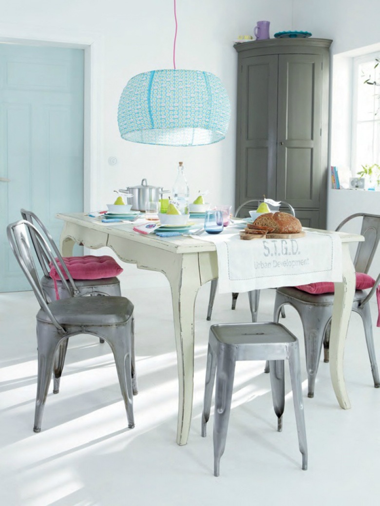 Prowansalski biały stół z drewna,szare krzesła i stołki industrialne z różowymi siedziskami,szara szafa i niebieska lampa nad stołem (27714)