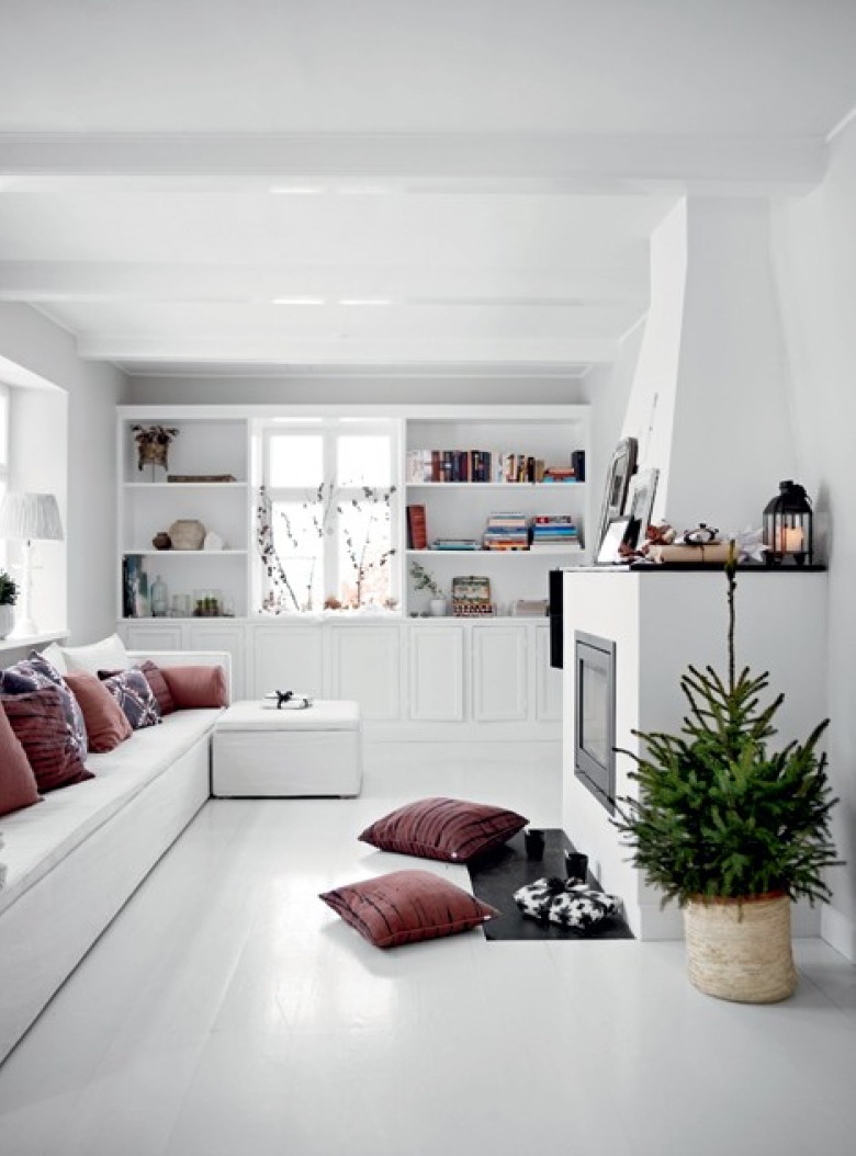 Biały salon jkominkowy z zabudowanymi siedziskami z poduchami i pufą,biała podłoga i półki wokół okna i malinowo-wrzosowe poduszki dekoracyjne (27407)