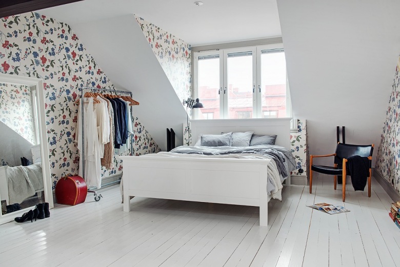 Białe łóżko,stojący metalowy wieszak na kółkach,drewniane ciemnobrązowe belki w sypialni,kwiatowa tapeta ścienna w niebiesko-czerwonym kolorze, duże białe lustro stojące (25941)