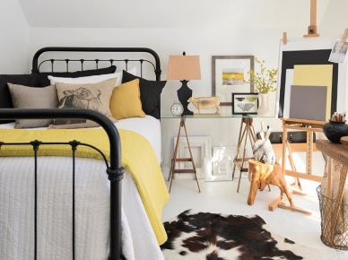 Kute czarne łóżko,stół na drewnianych kozłach ze szklanym blatem,czarno-zółte dekoracje łóżka,czarna lampa z bezowym abażurem i dywan z bydlęcej skóry na białej podłodze (26163)