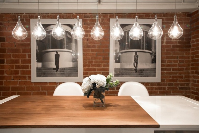 Żarówki na kablach nad drewnianym stołem w jadalni ze ścianą z czerwonej cegły i czarno-białymi fotografiami (26724)