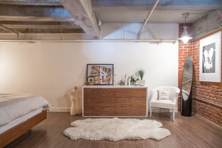 Biały dywan futrzak,białe ściany,przemysłowy sufit z betonowymi belkami,nowoczesna biało-drewniana komoda,stylowy biały fotelik i ściana z czerwonej cegły w sypialni (26732)