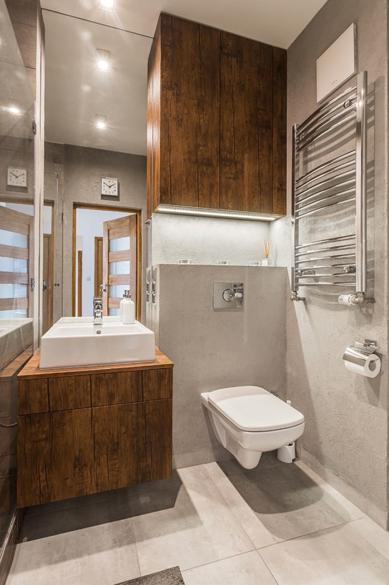 Łazienka w nowoczesnym stylu, w której zestawiono ze sobą szarość oraz ciemne drewno. Wysokie lustro nad umywalką...