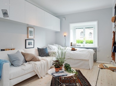 Białe wiszące szafki,łóżko przy oknie i biała sofa, okagłymetalowy stolik z tacą i etniczny brązowy dywan w salonie połączonym z sypialnią (25910)