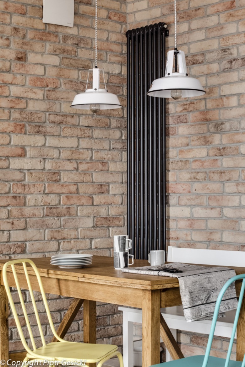 Surowe cegły na ścianie w jadalni wyraźnie eksponują szczególnie kolorowe krzesła przy drewnianym stole. Białe industrialne lampy wiszące wpisują się w ogólny charakter wnętrza. Ciekawie rozwiązano kwestię ogrzewania, montując na ścianie pionowy grzejnik w czarnym...