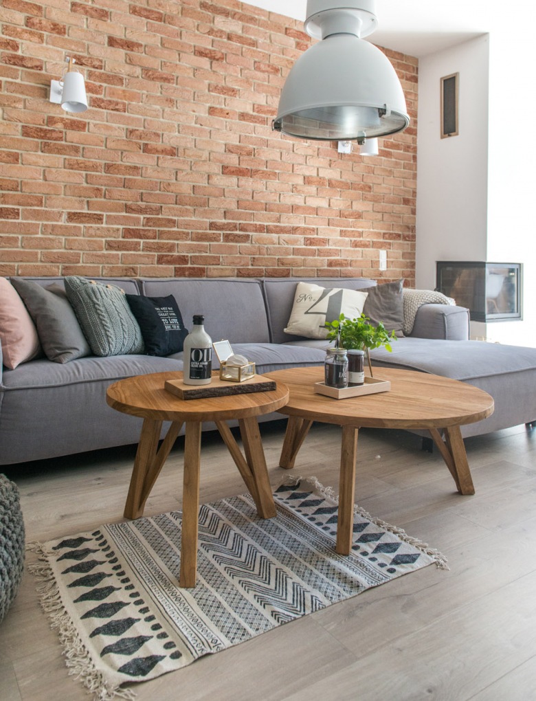 Drewno oraz czerwone cegły to wyjątkowe materiały, które tworzą przytulny klimat w salonie. Dla kontrastu znajduje się tu szara sofa o prostym kształcie. Kominek potęguje wrażenie...