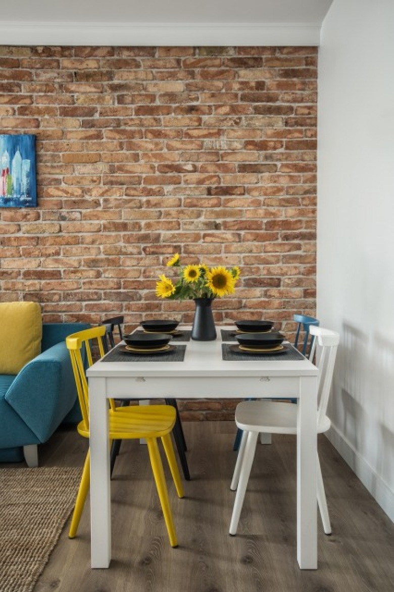 Żółty kolor krzesła wyróżnia się na tle ceglastej ściany. Bardzo wdzięcznie nawiązują do niego dekoracje z małych...