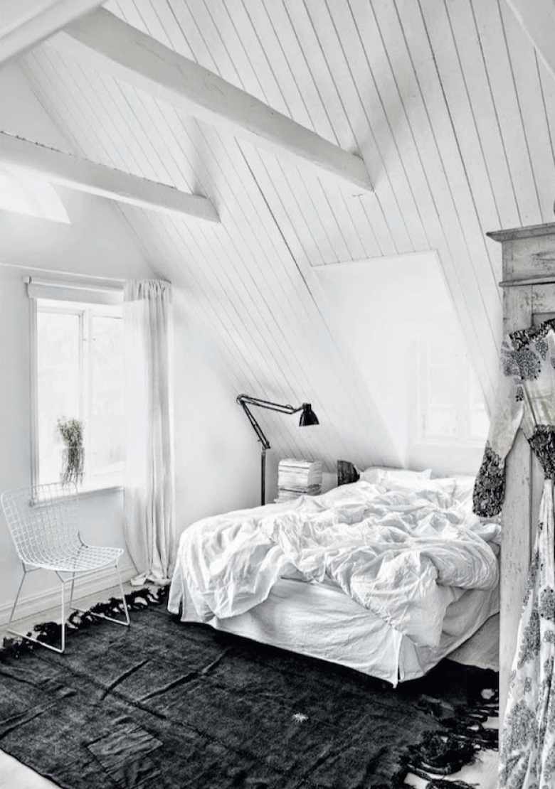 Biała sypialnia ze skośnymi ścianami z białymi belkami i czarnym dywanem z frędzlami na podłodze (23647)