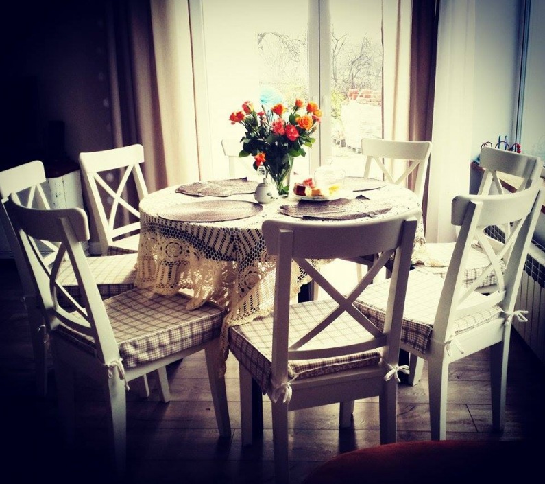 Przyjemna aranżacja jadalni w rustykalnym stylu z okrągłym stołem. Wzorzyste poduszki na krzesłach dekorują przestrzeń,...