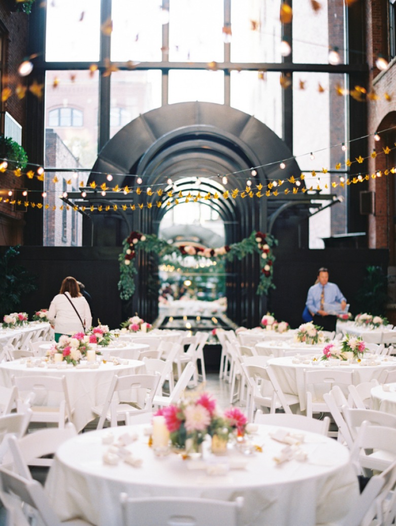 Pomysł na dekoracje sali na wesele. Klasyczne białe obrusy i krzesła, stoły ozdobione bukietami różowych kwiatów, a całość oświetlona girlandami świetlnymi, które znacznie ocieplają surowe...