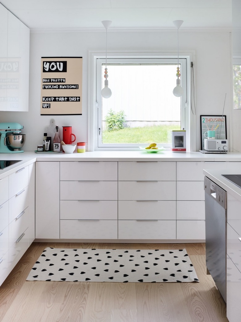 Niemal zupełnie białą przestrzeń w kuchni nieco ożywiają zastosowane dodatki. Czarne akcenty na dywanie czy blacie oraz...