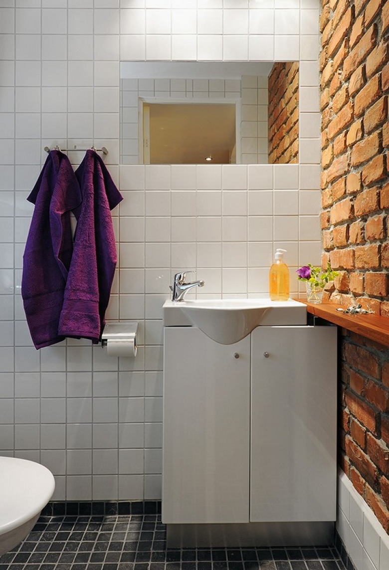 Surowa łazienka, białe kafelki i ciemna podłoga ożywione dzięki ścianie z czerwonej cegły.