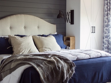 Żyrandol w sypialni i drewniane belki to bardzo odważny duet. Do tego eleganckie łóżko z bezowym zagłówkiem i poduchami idealnie wpasowuje się w sielski klimat. Umocowanie lampy z wysięgnikiem do bocznej ściany białej szafy to dobry pomysł na małe...