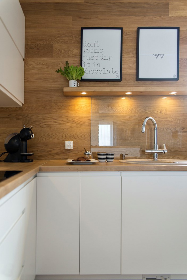 Drewniana ściana w kuchni pięknie komponuje się z dominującą bielą. Gładkie fronty szafek podkreślają prostotę stylu...