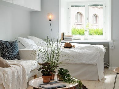 Białe wiszące szafki,łóżko przy oknie i biała sofa, okagłymetalowy stolik z tacą i etniczny brązowy dywan w salonie połączonym z sypialnią (25911)