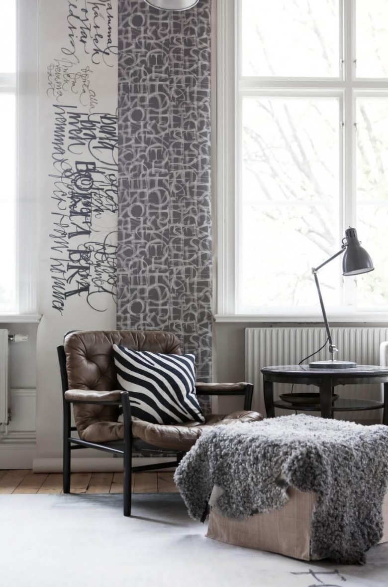 Graficzne wzory tapet w aranzacji skandynawskiego salonu ,czarny drewniany fotel ze skórzana tapicerką,kwadatowy puf z szarym futrzakiem i okragły czarny stolik kawowy (25992)