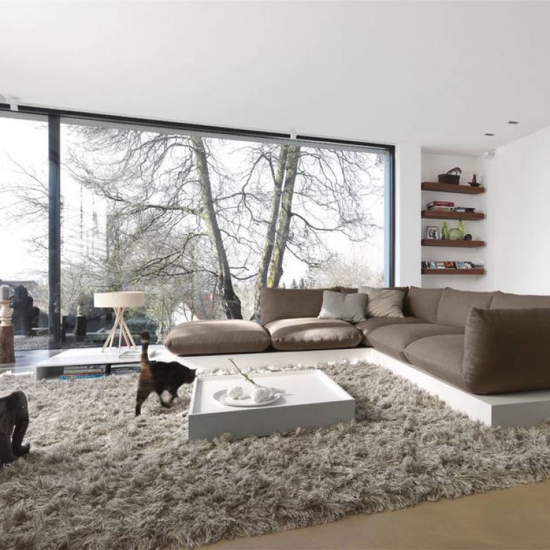 Panoramiczne okna z widokiem na drzewa,szara sofa narożnik na białym postumencie,szary dywan futrzak,biały niski nowoczesny stolik taca,drewniane półki w białej wnęce salonu (48033)