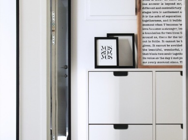 Wąskie czarne lustro na białej ścianie,biale szafki wiszące na buty,czarna półka,grafiki i typografie w czarnych i drewnianych ramkach w dekoracji przedpokoju (25758)