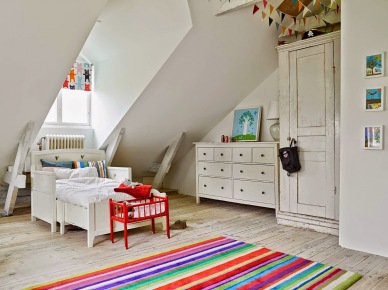 Białe skrzyniowe łóżko dziecięce,biała komoda z szufladami,kolorowa girlanda z proporczykami i kolorowy dywanik w paski w dziecięcym pokoju (26009)