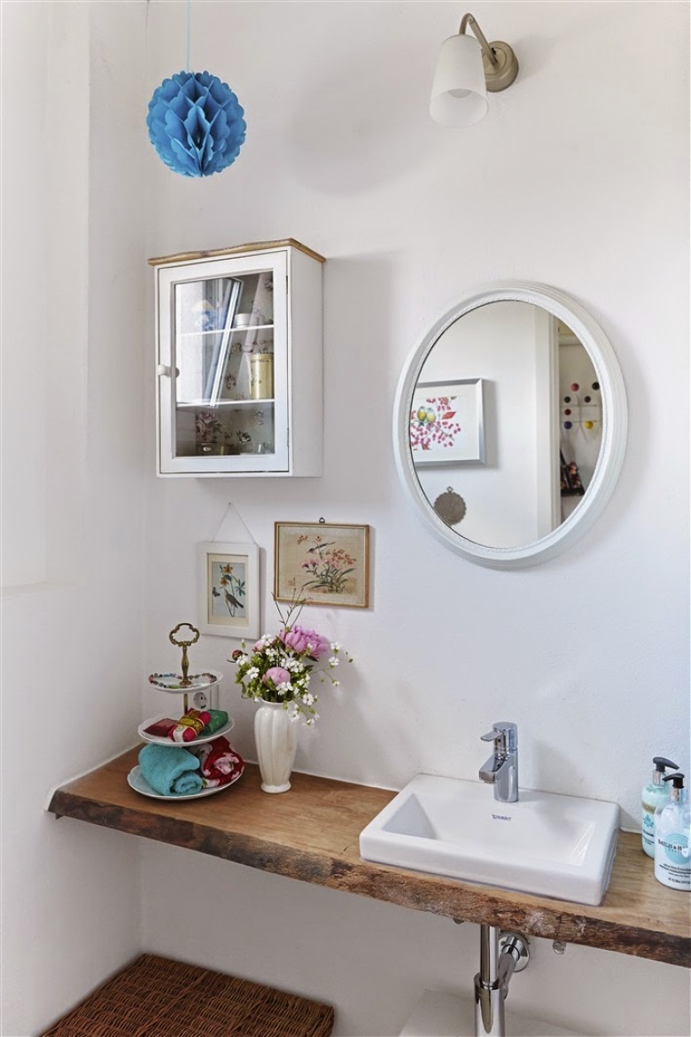 Białą łazienkę urozmaicają dodatki w żywych kolorach oraz w szczególności drewniany blat pod umywalką. Oryginalne akcesoria i ozdoby takie, jak kwiaty w wazonie przemieniają typowo łazienkowy klimat w bardziej 