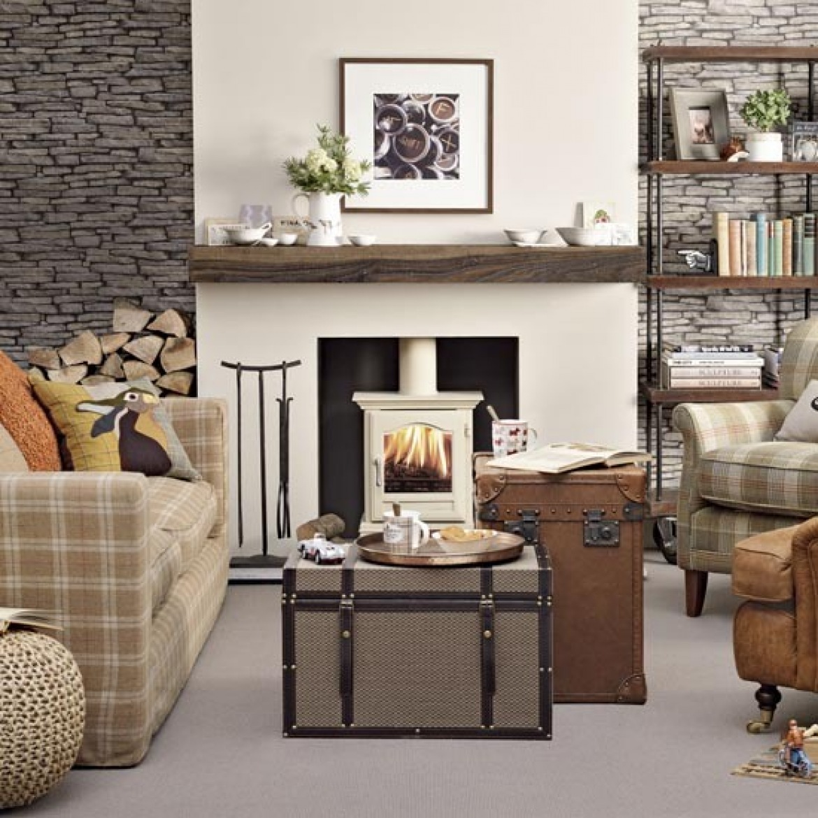 Kamień łupkowy na ścianie w salonie z białym kominkiem z drewnianą półką,kufry skrzynie w roli stolków i kraciaste sofy i fotele (26101)