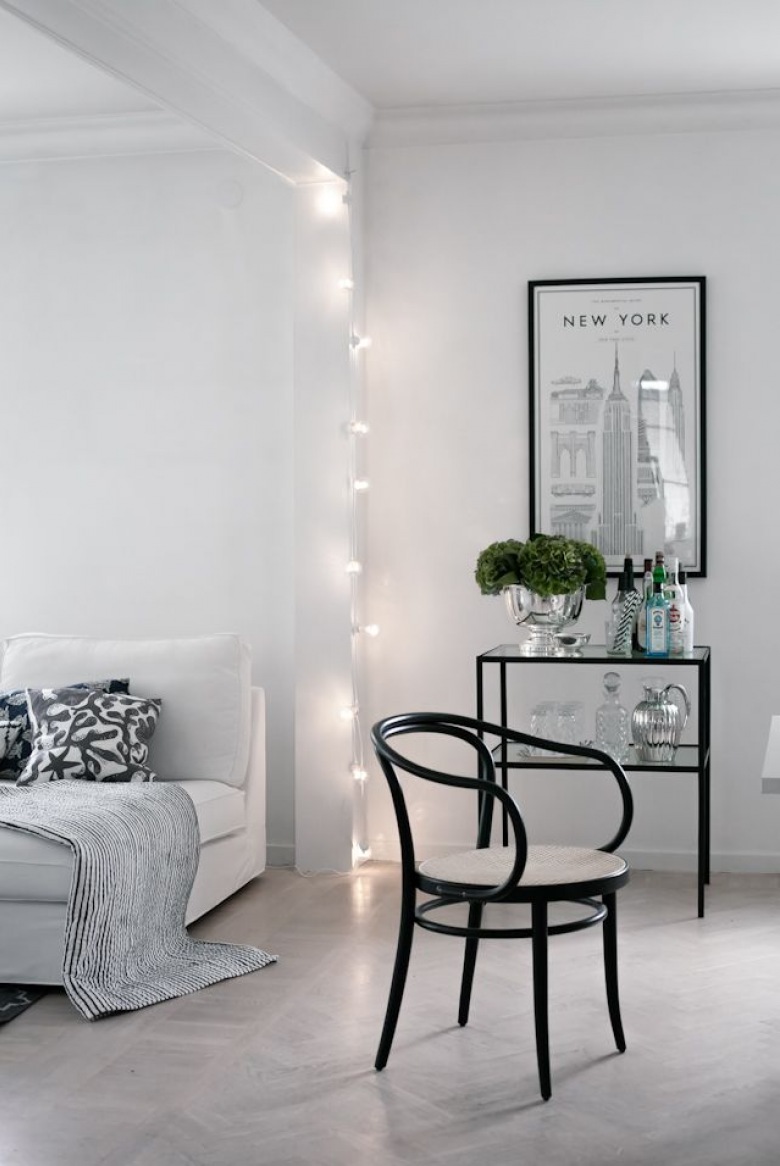 Surowy salon w bieli z czarnymi elementami w postaci krzesła i konsolki wydaje się być bardzo minimalistyczny, ale to właśnie girlanda ze światełkami wprowadza tu przytulny...