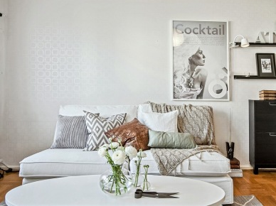 Stonowane pastelowe poduszki i pledy na białej sofie,czarna komoda i czarna girlanda z żarówek,srebrna tapeta na ścianie w salonie (28483)