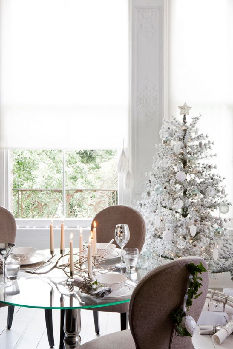 Biała i oszroniona dekoracja zielonej choinki przy okrągłym szklanym stole i tapicerowanymi krzesłami w jadalni z białymi roletami na oknach (27423)