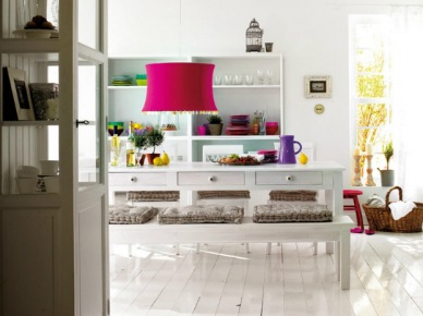 Biały prostokątny stół z szufladami,biała ławka z szarymi siedziskami,białe deski na podłodze i lampa w kolorze fuksji nad stołem (27713)