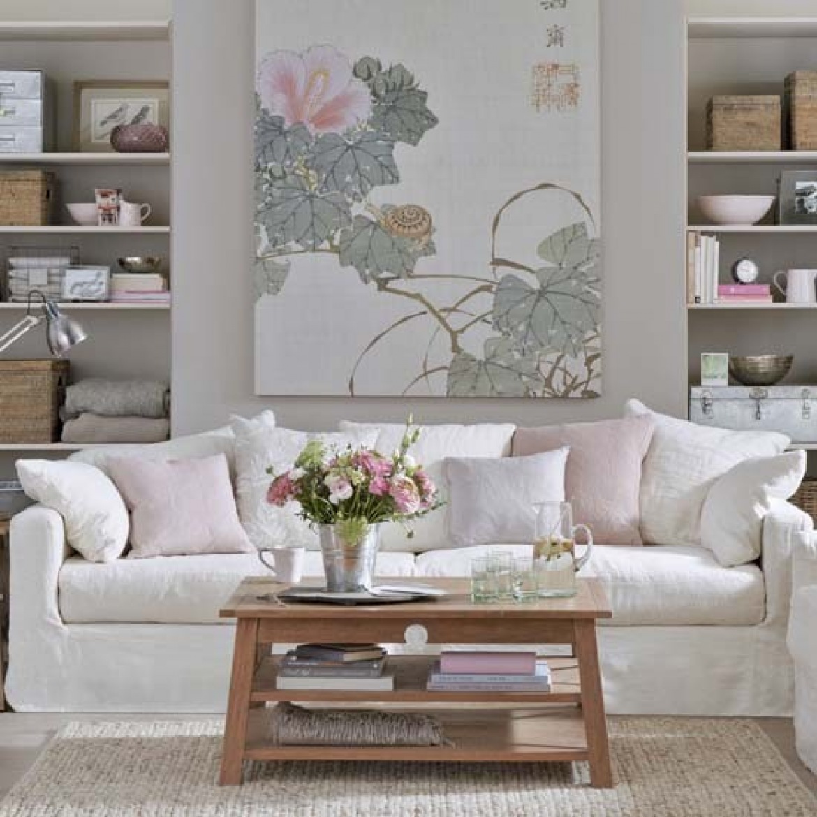 Drewniany tradycyjny stolik kawowy,szaro-różowy obraz z motywem roślin nym,jasnoszare ściany w wbudowanymi białymi pólkami i biała sofa na tkanym dywanie w salonie (26110)